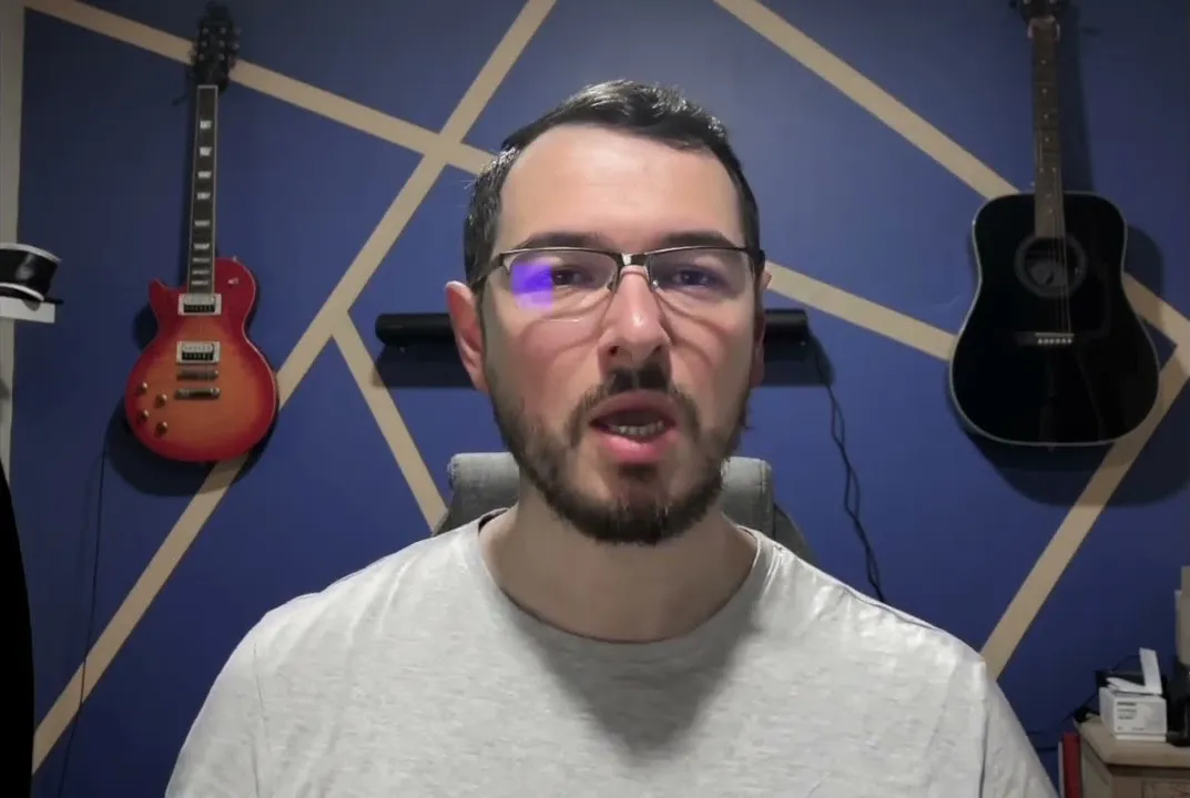 a screenshot of a Youtube video : a guy with short hair and a pair of glasses is talking to the camera with many guitars on the background